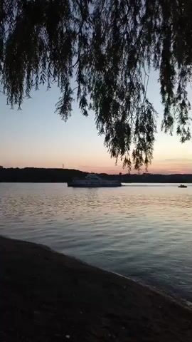 A sunset scene over a river. A ferry boat travels across the water, and its wake is visible. Tree branches hang down from the top of the frame, obscuring parts of the sky. The water is calm, with small ripples. The shore in the foreground is dark and indistinct. The sky is a soft gradient of blue and pink.
