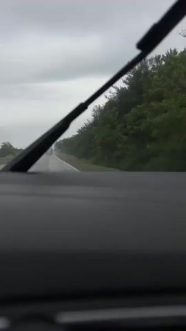A first-person perspective video shows a view from inside a vehicle traveling on a wet road on a cloudy day. The windshield wipers are intermittently visible, sweeping across the lower portion of the frame. A line of trees runs along the right side of the road, and in the distance, several trucks are visible on the highway. A circular traffic sign with a red border and a red line through a truck symbol is briefly visible on the right side of the road. The road surface appears wet and reflective.