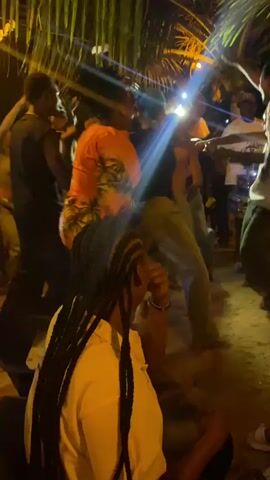 The video shows a group of people dancing outdoors at night under palm trees. They are dressed in casual clothing and appear to be enjoying themselves. The scene is illuminated by bright spotlights and the music is audible. Some people are holding up their phones, likely recording the event. There are tables and other equipment in the background, suggesting a possible performance or organized event.