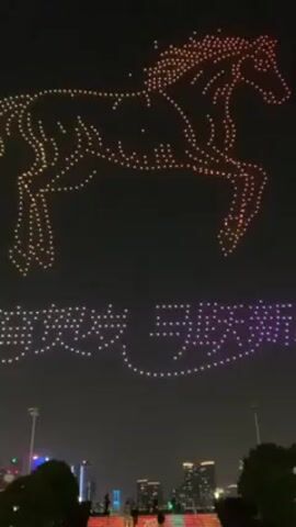 The video shows a drone light show in an urban environment. In the night sky, a horse figure comprised of numerous lit drones glows with orange and yellow hues. Below, another formation of drones creates a message in purple and white lights. Several individuals can be seen on stairs in the foreground, presumably watching the display. The urban backdrop includes buildings and greenery, indicating a city park or similar public space.  A single loud boom, presumably from a firework, can be heard in the middle of the video.