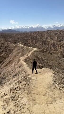A man in a black outfit is walking away on a dirt path. The path is on a ridge with a steep drop-off on either side. The landscape is a dry, hilly area with eroded rock formations. In the distance, there are snow-capped mountains under a clear blue sky with a few small clouds.