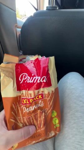 The video shows the interior of a car. A hand holds a bag of "Prima XXL Pack Peanuts" in the back seat. The camera pans to show the driver, a man wearing a white shirt and baseball cap, and then shows the view out the window, revealing a building with "KRALJI" on the front. The car is moving. The interior is mostly black and grey.