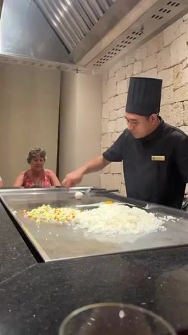 A chef in a black uniform and chef's hat stands behind a large flat-top grill. In front of him, the grill is divided into sections. One section contains chopped vegetables, another has a pile of white rice, and a third has two eggs, one whole and one cracked. The chef is using a spatula and fork to manipulate the food on the grill, specifically cracking the second egg. To the left of the grill, a woman in a floral-print dress is seated, looking at the chef. The background shows a stone wall and a large metal hood above the grill.