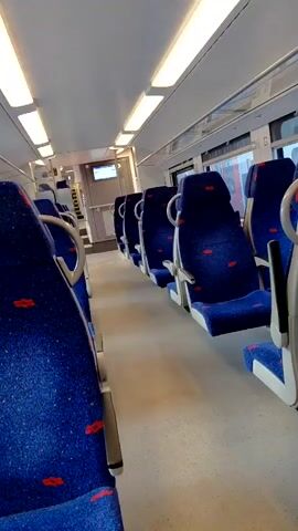 The video shows the interior of a train carriage. The seats are upholstered in blue fabric with a red pattern and are arranged in rows on either side of a central aisle. The carriage is well-lit by fluorescent strip lights on the ceiling. Windows run along one side of the carriage, showing a blurred view of the outside. The camera pans slowly, giving a clear view of the empty seats and the general layout of the carriage. The environment appears clean and well-maintained. There are no people visible in the carriage.