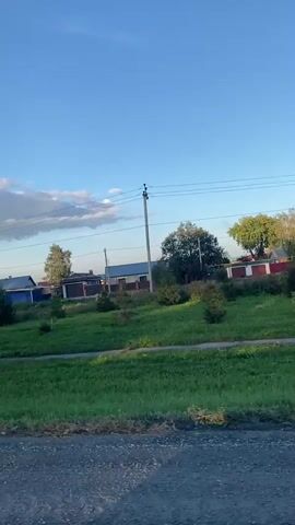The video shows a view from a moving vehicle, looking towards a rural residential area under a clear blue sky with some scattered clouds. In the foreground, there is a rough, greyish surface, likely a road. Beyond that, a strip of green grass with some small evergreen trees is visible. In the mid-ground, there are several houses with various roof colors, including blue, white, and brown, separated by fences, some of which are blue. Power lines and a utility pole are also present in the scene. The overall impression is that of a quiet, daytime scene in a village or suburban area.