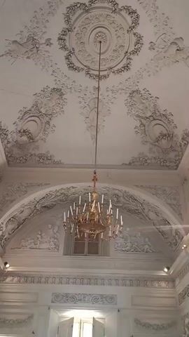 The video shows the ornate ceiling and a large, gold chandelier hanging in a grand room, likely a palace or historic building. The ceiling is elaborately decorated with intricate plasterwork, featuring floral motifs, swags, and possibly mythological figures in relief. The chandelier is made of brass or gilded metal and holds numerous white candle-like light fixtures.

Below the ceiling and behind the chandelier, there is a recessed area with a window, and further relief sculptures depicting human figures can be seen. The walls also feature detailed decorative moldings and architectural elements. The overall impression is one of opulence and historical grandeur, characteristic of Italian Baroque or Rococo architecture.

The lighting suggests it might be daytime, but the artificial lights from the chandelier are also prominent. There is no visible text in the video. The setting appears to be a well-maintained interior, possibly a museum or preserved historical site in Colorno, Italy. The focus is on the architectural and decorative artistry of the room.