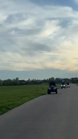 Three people are riding ATVs on a paved road. The ATVs are moving toward the camera. The sky is cloudy. There are fields and houses in the background.
