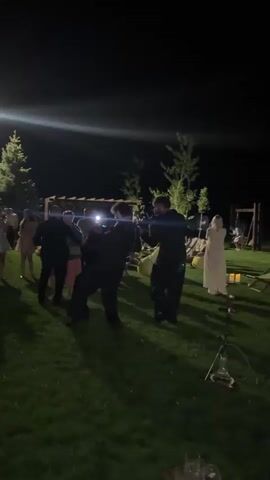 The video shows an outdoor nighttime gathering on a grassy lawn. Several people are visible, some dancing and interacting. In the center, two individuals appear to be performing or entertaining a group. One is playing a musical instrument, possibly a clarinet or a similar wind instrument, while the other stands nearby, possibly singing or participating in the performance. Other individuals are observing the performance or dancing in the background. The environment is dark, with artificial lighting illuminating the area. There are trees and some structures, possibly a pergola, visible in the background. The overall scene suggests a social event like a party or a celebration.