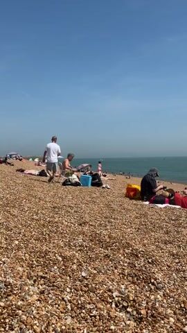 The video shows a pebble beach with several people sunbathing and relaxing. In the foreground, two men are standing and sitting near a blue cooler. A woman is lying down on the pebbles beside them. To the right, another person is sitting near an orange bag.  In the background, more people are sunbathing and swimming in the ocean. The sky is clear and blue. The overall scene is a sunny day at the beach.