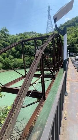 The video shows a view from a bridge overlooking a turquoise river. The bridge has a pedestrian walkway on the left, separated from the road by a metal railing. The road appears to have two lanes and is in good condition. In the distance, there are mountains covered in lush green trees. The sky is blue with scattered white clouds. A tall power pylon is visible in the background. A white van is driving on the bridge. On the right side of the bridge, there is a modern building with large windows and stone accents. The river flows under the bridge.