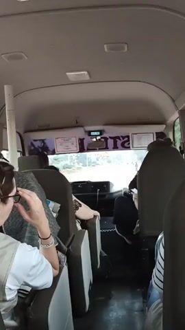 The video shows the interior of a van-like vehicle from a rear passenger perspective. Several people are seated inside, and one person in the foreground is wearing a white top and has bracelets on their wrist. Through the front windshield, a street scene is visible with people walking. The dashboard includes a digital clock showing 5:48, various stickers, and a rearview mirror. The vehicle appears to be in motion.