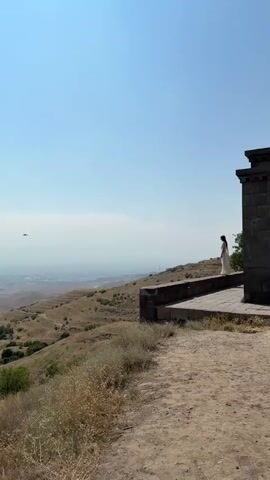 A vertical, medium shot captures a woman standing on a stone terrace overlooking a vast, hazy landscape. The woman is wearing a long, white dress and is seen from the side, facing away from the camera. She is positioned to the left of a dark, stone structure, the corner of which is visible on the right side of the frame. The terrace appears to be part of an ancient or historical site, with stone railings and weathered stone work. Beyond the terrace, the ground slopes downwards, covered in dry, yellowed grass and sparse vegetation. The distant landscape is a panorama of undulating hills and what appears to be a city or town shrouded in haze. The sky above is a clear, bright blue. A faint, dark object, possibly an airplane, is visible in the upper left quadrant of the sky. The overall impression is one of serene contemplation and vastness.