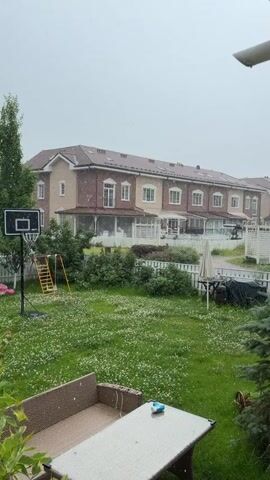 The video shows a suburban backyard with townhouses in the background. It is raining, with visible raindrops in the air. The yard is covered in green grass with white flowers. A basketball hoop and a swing set are visible in the yard. There is a picnic table and chairs in the foreground. A fence separates the yard from the townhouses. The sky is overcast.