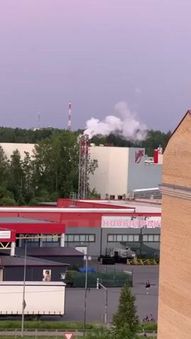 The video captures an outdoor industrial scene. In the background, a large white building with a red logo is visible, with white smoke emanating from a tall, red and white striped smokestack. Trees surround the industrial area. In the middle ground, a smaller building with a red roof is present, featuring a sign with Russian text. A white truck is parked in front of this building. The foreground shows a paved area where two figures are walking, one with a dark shirt and shorts, and another with lighter clothing. Two pink bicycles are parked near the figures. The sky is overcast with a purplish hue. A brick building is partially visible on the right edge of the frame.