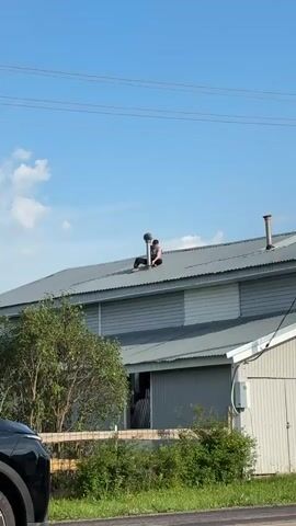 A person is sitting on the roof of a building next to a chimney vent. The person is wearing dark pants and a dark tank top. The person's hands are on the chimney vent. The roof is made of corrugated metal. There are trees and bushes in front of the building. The sky is blue with some clouds.