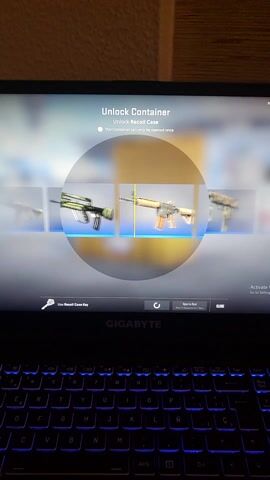 A laptop screen displays a video game interface, primarily focused on an item unlocking mechanism. The screen is oriented vertically in the image.

In the initial moments, the screen shows a central circular area where various weapon skins are rapidly cycling, mimicking a "case opening" animation. To the left of this circle, text reads "Unlock Container" and "Unlock Recoil Case." Below this, there's a small circle with a dot, indicating a selected option, next to the text "This container can only be opened once." At the bottom right of the screen, a key icon is present with the text "Use Recoil Case Key." Above this, the word "GIGABYTE" is visible, likely a branding for the laptop or game component. In the top right corner, a "CLOSE" button is displayed.

As the video progresses, the animation stops, and a specific weapon skin is revealed in the center of the screen. This skin is identified as "P90 | Vent Rush" with "The Recoil Collection" displayed underneath. A descriptive text accompanies the item: "Easily recognizable for its chrome finish, this P90 is a weapon for those on the move due to its low weight. It has been custom painted with a detailed, metallic graphic over a base of bullet casings, tubes, and fire." Below this, a question reads, "Do you know how just you were going?". The background behind the weapon shows what appears to be an airport or airplane scene with hints of white and red.

To the right of the screen, a portion of the laptop's keyboard is visible. The keys are illuminated with a blue backlight, and white legends are printed on the dark keycaps. The "GIGABYTE" logo is also present on the laptop's body, below the screen. The laptop appears to be resting on a light-colored surface, possibly a desk or floor, with a textured wall visible to the left of the screen.