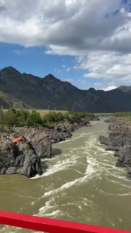 The video shows a wide, swiftly flowing river bordered by rocky terrain and a distant mountain range under a partly cloudy sky. On the rocky bank, a small group of people gathers around a large, spread-out orange cloth. Some are dressed casually, while others wear more formal attire. The camera angle suggests the video is taken from a bridge with a red railing visible in the foreground. The scene appears to be a riverside gathering or event.