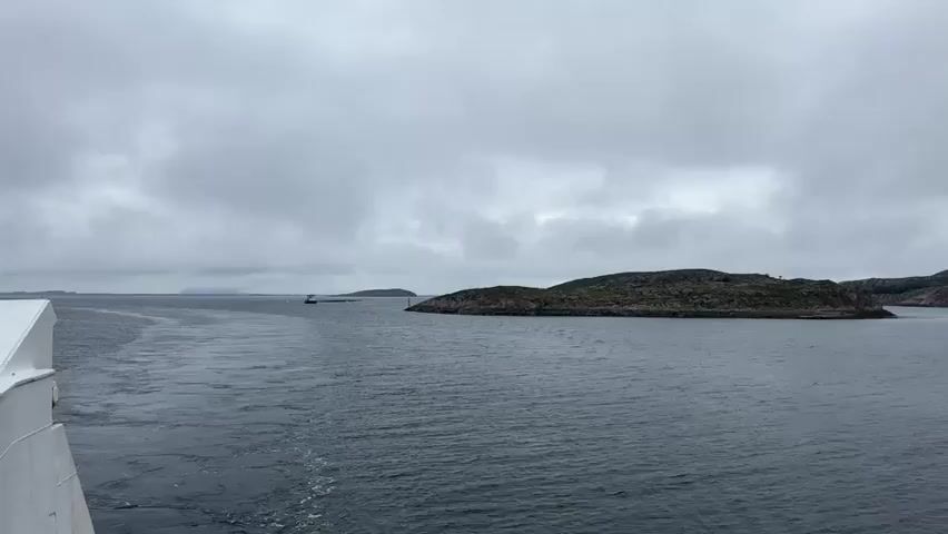 A wide shot of a body of water with several small, rocky islands covered in green vegetation. The sky is overcast with gray clouds. On the left side of the screen, the wake of a boat is visible. In the distance, there are a few small buildings scattered across the islands. The camera pans slowly to the right, revealing more islands and the open sea. The video is taken from the perspective of someone on a moving boat. There are no people visible in the video.