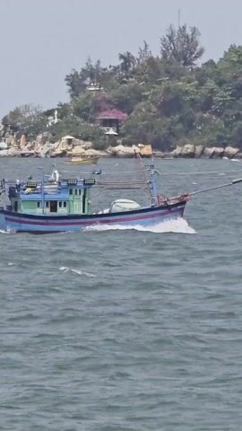 A blue and green fishing boat moves across choppy water, heading toward the viewer. The boat has a cabin in the middle, poles and ropes for fishing, and a red stripe near the top. In the background, a rocky shoreline with trees, buildings, and what appears to be a pink gazebo is visible.
