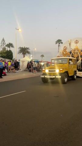 The video shows a yellow Toyota Land Cruiser pulling a large float along a paved street, with crowds of people on the left. The Land Cruiser is driving with its lights on, pulling a large, ornate float decorated in gold and white. People stand along the sidewalk, watching the float pass. The surrounding environment includes palm trees and street lights, indicating an urban setting. The lighting suggests it is either early morning or late afternoon.