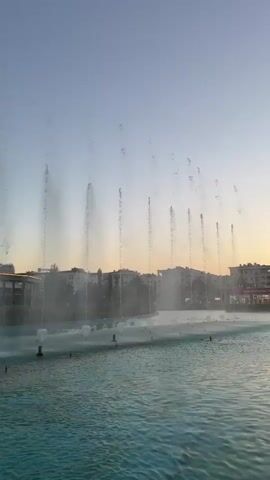 The video captures an outdoor scene at dusk. A large, turquoise body of water dominates the foreground, with multiple fountains spraying vertical streams of water upwards. These fountains are illuminated from below with yellow and blue lights, creating a vibrant effect. In the background, several modern buildings of varying heights are visible. The tallest building is a sleek, glass skyscraper with a darker section at the top. Adjacent to it is another, slightly shorter skyscraper. In front of these, a curved, lower building with a lit-up, colorful band around its upper section is also visible. The sky above is a clear gradient of pale blue to a warm orange near the horizon, indicating the setting sun. The overall atmosphere is serene and visually appealing, with the interplay of water, light, and architecture.