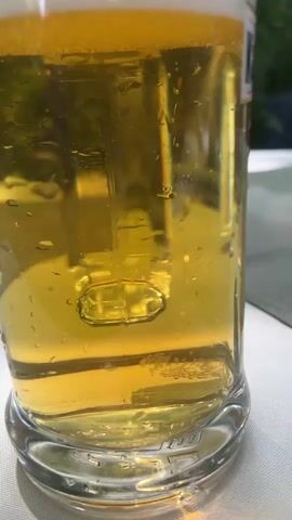 A close-up video begins showing a glass of beer with foam on top. The camera then pans to the right, revealing a scenic view of a waterfall cascading down a rocky, moss-covered cliff face. The water flows into a clear pool below. Trees and greenery surround the waterfall. The video appears to be shot outdoors, possibly from a table where the beer is placed.
