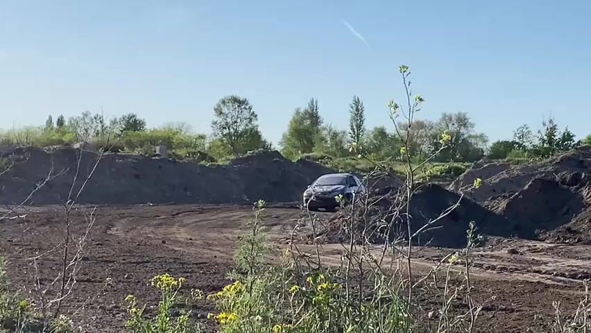 A blue hatchback rally car with the number "211" is driving quickly on a dirt track. The car kicks up a significant amount of dust as it drives past a clump of green and yellow weeds. The surrounding environment includes dirt mounds and greenery in the background, with a clear blue sky above. In the far background, there are buildings and construction equipment visible.
