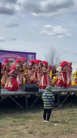A group of young women dressed in colorful traditional costumes are performing a dance on a stage. The costumes are predominantly red, gold, and yellow, and the dancers wear matching headpieces. The stage is a simple, metal platform with black speakers on either side. A banner with a Russian inscription that includes the word “festival” is visible in the background. Two young boys stand in front of the stage watching the performance. One boy wears a blue and green striped jacket and the other wears a dark jacket with white patterns. The background features a grassy field with bare trees, and a cloudy sky. The video appears to have been taken outdoors on a windy day.