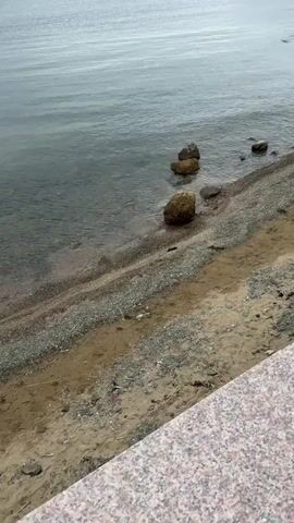 The video shows a view of a shoreline. In the foreground, there is a textured surface that appears to be a railing or ledge, possibly made of granite. The middle ground shows a beach with sand and small stones. Several larger rocks are visible along the edge of the water. The background consists of the body of water, which appears to be a lake or the sea, with calm, rippling surface. The water is a muted gray-blue color. The sky is not visible in the frame. The lighting suggests an overcast or cloudy day. There are no people visible in the video. The overall scene is natural and quiet, focusing on the transition between land and water.