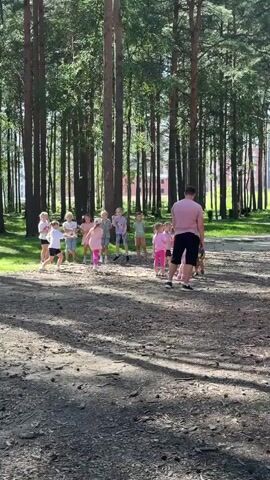 A group of children and one adult male are gathered in a clearing surrounded by tall pine trees. The children, appearing to be between the ages of 5 and 10, are dressed in casual, colorful clothing, with many wearing pink. They are arranged in a loose semi-circle facing the adult male, who is wearing a pink t-shirt and dark shorts. The children are actively engaged, with some raising their arms, jumping, or appearing to be dancing. The ground is covered with dirt and fallen pine needles, and dappled sunlight filters through the trees, creating shadows on the ground. In the background, buildings are visible through the trees. The overall impression is of an outdoor recreational activity or a children's event taking place in a wooded area.
