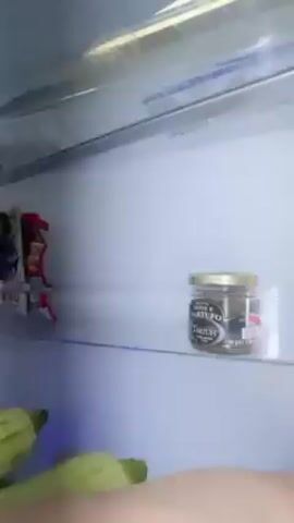A woman is filmed from inside a refrigerator. The refrigerator is mostly white with glass shelves and is rather empty. On the top shelf, there is a bottle, some small packets, and a small jar. On the bottom shelf, there are several whole zucchinis, a bottle, and a blue container. The woman reaches into the fridge, grabs two zucchinis, and holds them up. She is wearing a light blue T-shirt and has brown hair. Behind her is a window covered with a white curtain.
