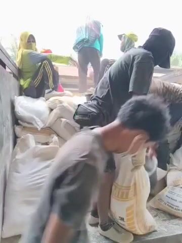 The video shows a group of people loading and unloading a truck filled with large sacks. Several individuals, some wearing headscarves, are visible in the back of the truck. One person in the foreground, wearing a dark t-shirt and shorts, is seen handling a white sack. Another person in a green top and headscarf is sitting on the edge of the truck bed. There are stacks of black buckets and many white sacks are piled up. The environment appears to be outdoors, with some vegetation and buildings visible in the background. The overall impression is of a cooperative effort to move goods.