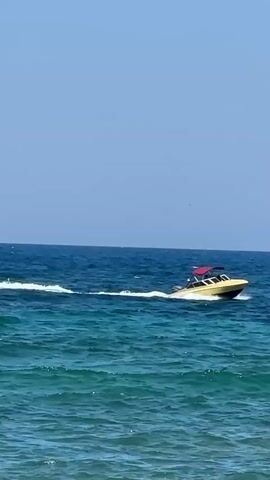 A yellow and red speedboat speeds across turquoise ocean water under a clear blue sky. The boat leaves a white wake behind it as it moves from right to left. The water is choppy with small waves.