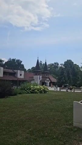 The video captures a building with two prominent pointed towers, resembling a whimsical castle or European-style architecture. Lush green trees and a well-maintained lawn surround the structure, suggesting a park or resort setting. In the foreground, a grassy area with some bushes and a concrete structure can be seen. In the mid-ground, in front of the building, there are several people. Some are standing and conversing, while others appear to be interacting with bicycles parked nearby. A few individuals are also seated on a bench in the background. The sky above is partly cloudy, indicating daytime. The overall impression is one of a pleasant, possibly tourist-oriented location, with a relaxed atmosphere.