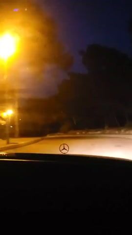 The camera is inside a vehicle, likely a Mercedes-Benz, judging by the logo visible on the dashboard, during a nighttime drive. The road ahead is illuminated by the vehicle's headlights, showing a winding path bordered by dark trees. Streetlights are also visible, casting a yellow glow on the road and surroundings. The sky is dark blue with hints of clouds.  The overall scene is dimly lit, with the road and the trees forming dark silhouettes against the night sky. There is faint audio present, although the specific content is not clear.