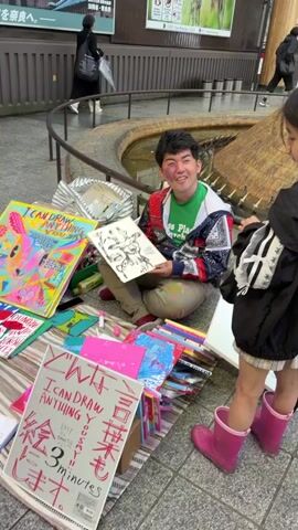 A medium shot shows a man sitting cross-legged on the ground, wearing a green t-shirt, a colorful patterned jacket, and tan pants, with some paint splatters. He holds up a black and white drawing of a cartoon character. To his left, several colorful drawings are displayed. To his right, a child in a black outfit and pink boots stands, partially visible. A white sign with red Japanese text and "I CAN DRAW ANYTHING YOU SAY" is in the foreground. In the background, there's a water feature, a curved railing, and a woman walking with an umbrella. A large building with a green banner is behind the railing.
