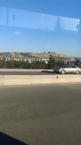 This media file captures a view from a moving vehicle, likely a bus or car, traveling along a highway. The scene unfolds under a clear, bright blue sky, suggesting a pleasant, sunny day. The time of day appears to be late morning or early afternoon, given the strong sunlight and the clear visibility of the landscape.

The primary subjects are vehicles on the highway. A dark-colored sedan is seen in the foreground of the first clip, followed by a large white semi-trailer truck with a white pickup truck behind it in the second clip. Further along the highway, in the third and fourth clips, a white sedan and a silver SUV are visible. The highway itself is paved and divided by a concrete barrier.

Beyond the highway, a sprawling city is visible on a gently sloping hill. Numerous buildings, ranging in height and architectural style, dot the landscape. Some appear to be residential apartment complexes, while others might be commercial structures. Trees and greenery are interspersed throughout the urban area and on the hillsides. In the distance, the terrain suggests a hilly or mountainous region.

The activity is straightforward: traffic moving along a highway with a city and its surroundings as a backdrop. There are no clear indications of emotions being conveyed, as the focus is on the scenery and the movement of vehicles. The overall impression is one of a typical journey on a well-traveled road, offering a glimpse of urban development against a natural landscape.

There is no visible text within the media file. The location context provided is Baku, Azerbaijan, which aligns with the visual cues of the urban development and the hilly terrain.