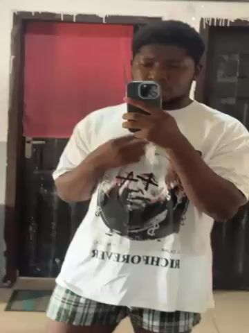A young man with dark skin and short curly dark hair is standing in front of a mirror, taking a selfie with his phone. He is wearing a white t-shirt with a graphic print and plaid pajama bottoms. Behind him is a closed dark-colored door with a silver handle and a red curtain or panel above it. The room appears to be a bedroom or similar personal space. He adjusts his t-shirt at the neck. The lighting in the room is moderate.