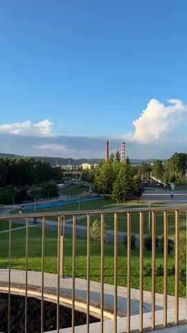 The video opens with a wide shot of a park-like area with green grass, trees, and a path. In the distance, there are industrial buildings with tall chimneys. The camera then pans to the right, showing more of the park and a bright blue sky with scattered clouds. The sun is setting, casting a warm glow on the scene. As the camera continues to pan, it reveals a walkway with a golden railing. The walkway curves and leads to a bridge-like structure with lamp posts. The video ends with a close-up of a person with red hair, partially visible in the foreground.