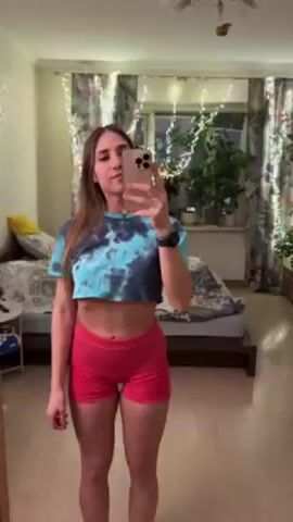 A woman stands in a bedroom, facing a mirror and holding up a phone to take a selfie video. She has long brown hair and is wearing a blue and black tie-dye crop top and red shorts. The room is decorated with curtains, string lights, plants, a bed, a dresser, and a TV. The woman makes small motions and adjustments while recording the video.
