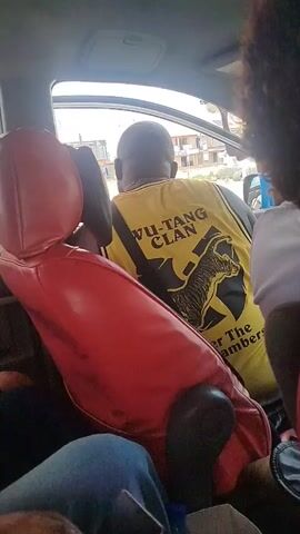 The video shows the interior of a vehicle from a passenger's perspective. In the foreground, red leather seats are visible. In the middle ground, a person wearing a yellow sleeveless shirt with "WU-TANG CLAN" printed on the back in black and yellow lettering is seated. The shirt also features a graphic of a claw holding a microphone and text that reads "ENTER THE CHAMBER". The person is facing away from the camera. Outside the vehicle's window, a street scene with buildings and other vehicles can be seen. The lighting suggests it is daytime.