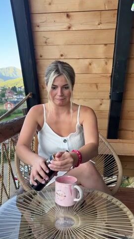 A woman with blonde hair is sitting in a chair on a balcony and pouring a dark liquid from a can into a pink mug. She is wearing a white tank top. The balcony is made of wood, and there is a table in front of her made of woven material. In the background, there are trees and houses on a hillside, with mountains beyond.