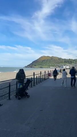 The video shows a seaside promenade on a sunny day with a blue sky and some clouds. A man in a dark jacket stands next to a baby stroller, looking at his phone. The promenade is bordered by a metal railing separating it from a pebble beach and the sea. In the background, a green hill or mountain rises, with some buildings visible at its base. Several people are walking along the promenade, moving away from the camera. The scene is bright, suggesting daytime.