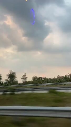 The video captures a cloudy dusk sky from the perspective of a moving vehicle. A ring of blue lights is visible in the sky, appearing to hover above a blurry landscape. The landscape features trees and some structures that are indistinct due to motion blur. A red light is also visible near the horizon, possibly the setting sun or another light source. The scene suggests a view from a highway, with metal barriers and grassy areas visible in the foreground. The overall mood is somewhat hazy and diffused due to the cloudy conditions and the motion of the camera. The location is identified as Wonosalam, Indonesia, and the time of day is dusk. There are no discernible people or clear text in the video. The blue ring of lights is the most prominent and unusual object in the scene.
