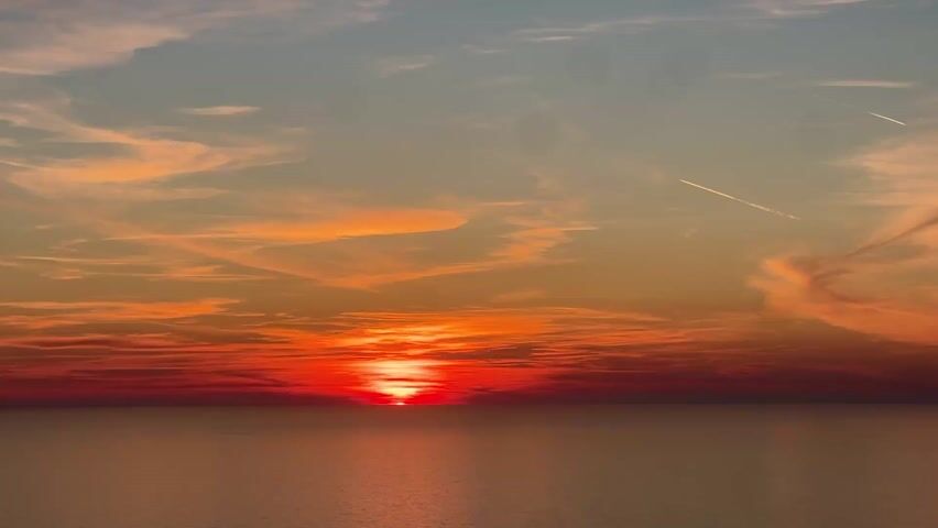 The video shows a vivid red sunset over a calm body of water. The sun sits right at the horizon, with its light reflecting off the water's surface. Above the horizon, the sky transitions from a deep red to orange, then to a lighter blue near the top of the frame. Wispy clouds are scattered across the sky. The sky also includes two contrails of airplanes. There are brief sounds of people in the video.