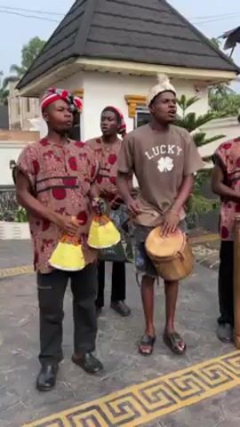 The video shows five men performing traditional music in front of a building. The men are dressed in African attire, with four of them wearing matching patterned tops and one wearing a casual "Lucky" t-shirt. They are playing traditional instruments, including a drum and bells, and singing. The building behind them has a black roof and gold trim. The video appears to be taken in an outdoor setting on a paved area. A portion of the frame shows a close-up of one of the men's patterned shirt.