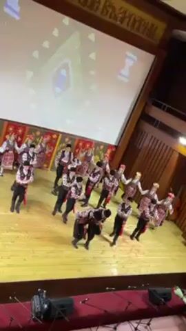 A high-angle shot captures a group of approximately 20 dancers on a wooden stage, performing what appears to be a traditional folk dance. The dancers are clad in colorful costumes with black pants and vests, adorned with red sashes. Some of the performers wear white head coverings with red or multi-colored accents. The dancers move in unison, stepping and extending their arms. Behind the dancers, a large projection screen displays geometric shapes. Above the screen, wooden paneling is inscribed with text in what appears to be Cyrillic script. The stage is bathed in light, accentuating the wooden surface. In the foreground, the lower portion of a red-carpeted audience section is visible, along with the corner of a wooden banister. The camera remains static.