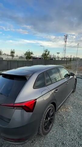 The video captures an outdoor scene on a clear day with a blue sky and scattered white clouds. It begins with a rear-left view of a gray station wagon car with black wheels, parked on a gravel surface. The camera slowly pans right, revealing a landscape behind the car. In the mid-ground, a dark fence line runs horizontally, behind which are green trees, a street light, and a large metal scaffolding-like structure. Further in the background, a tall metal cell phone tower with multiple antennas stands prominently. A pole with solar panels and a light fixture is also visible on the right side of the frame. The area to the far right appears to be open, undeveloped ground. No people are visible in the video.