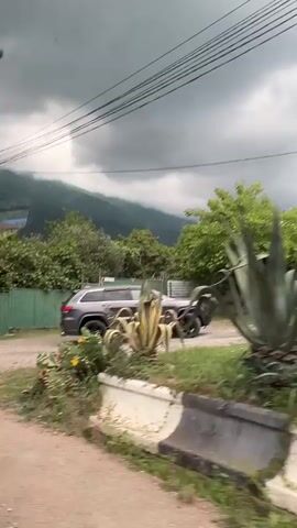 A dark grey SUV is seen driving on a dirt road. The SUV is turning right, entering a property through a gate. The gate is green and made of metal. Beyond the gate, there are trees and lush greenery. In the background, there are mountains covered in clouds and mist. There is a tall lamppost with power lines running across the frame. The sky is overcast with dark clouds, suggesting impending rain or a storm. There are some plants and flowers visible in the foreground. The overall impression is a rural or semi-rural setting with a dramatic, cloudy sky.