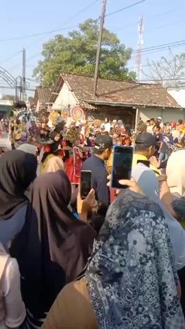 The video shows a crowd of people watching a parade or festival. Several individuals in colorful costumes, some with elaborate headdresses, are visible in the center of the frame. Many people in the crowd are holding up their phones to record the event. The background shows houses and trees under a clear sky. The overall atmosphere appears festive.